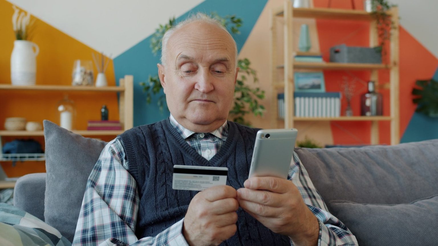 Imposter Scams Are Targeting Seniors: How to Spot the Warning Signs Before It’s Too Late Elderly man holding credit card and smartphone for online shopping.